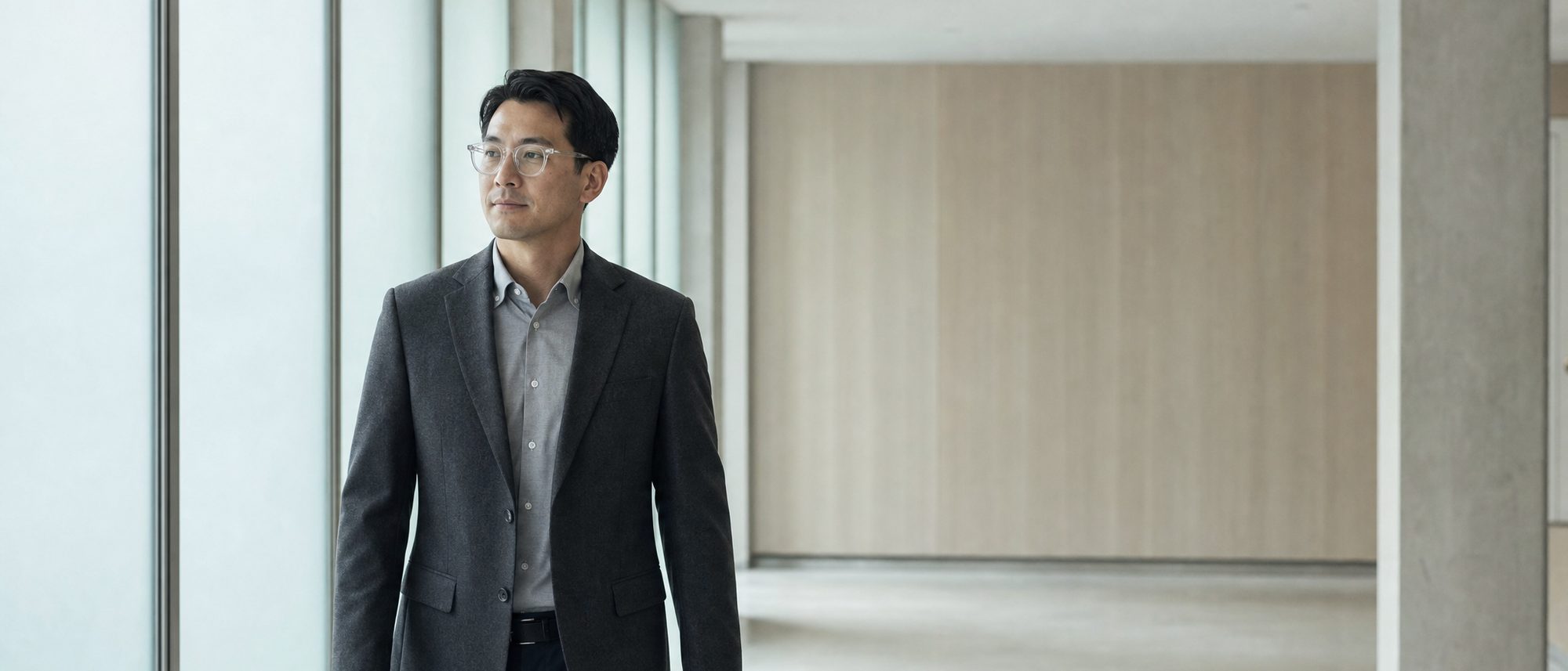밝은 실내 통로를 걷는 사람이 얇은 안경을 쓰고 있고 양손은 비어 있다